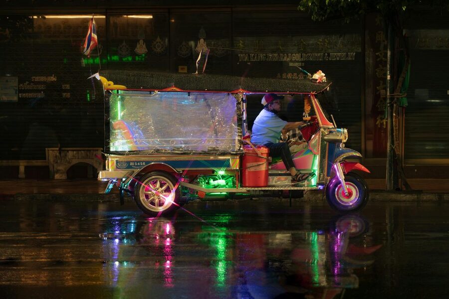 Tuk-tuk driving through rainy Bangkok street with neon reflections