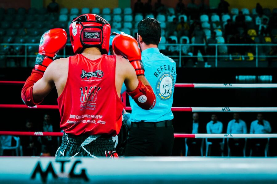 Rear view of a Muay Thai boxer in red gear facing the referee inside a boxing ring