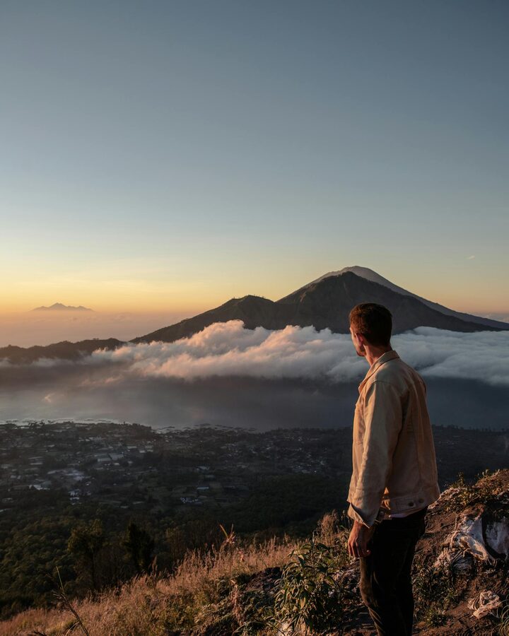 Person standing on a hillside overlooking Mount Batur in the morning light