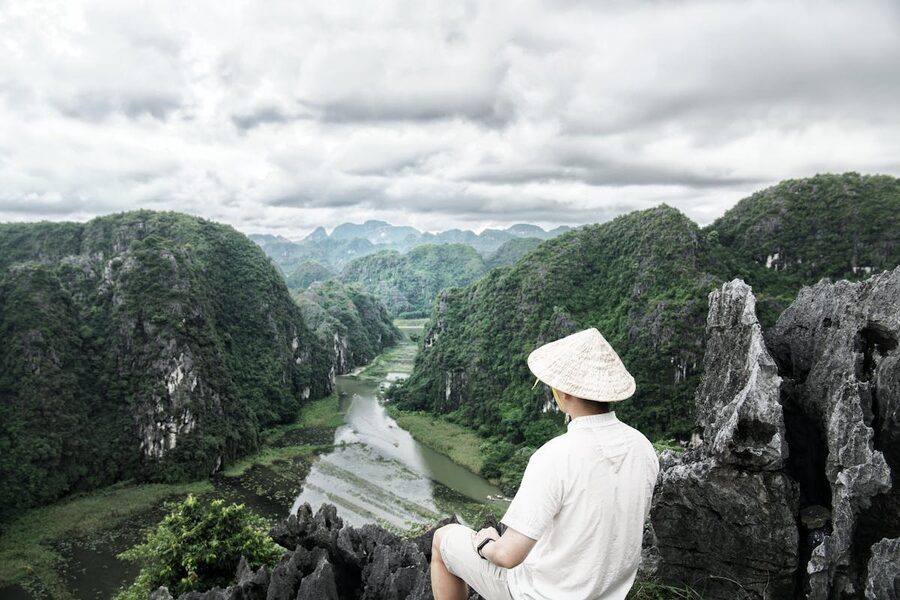 A man with a conical hat sits atop a cliff overlooking the lush Trang An valley in Ninh Binh