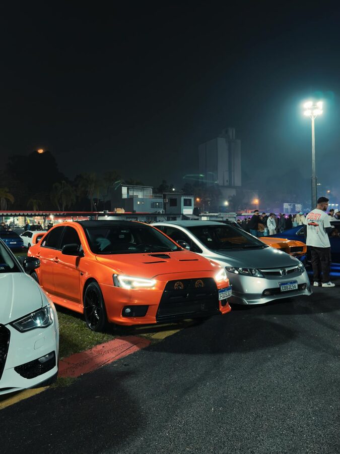 Bright orange sports sedan surrounded by other vehicles at a nighttime car meet