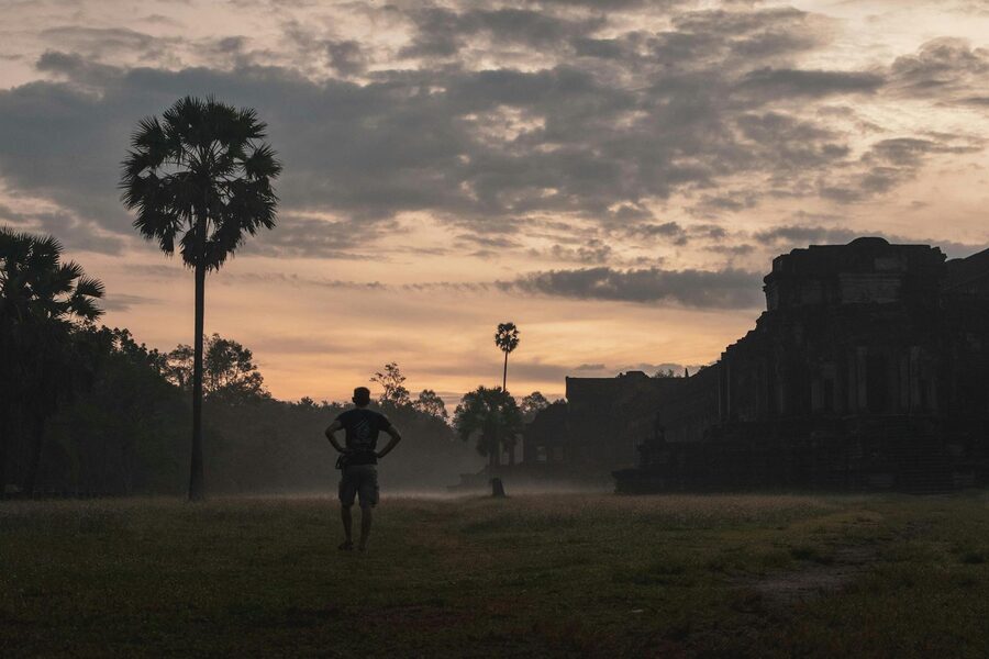 A man stands in silhouette near Angkor Wat at sunrise, surrounded by other visitors