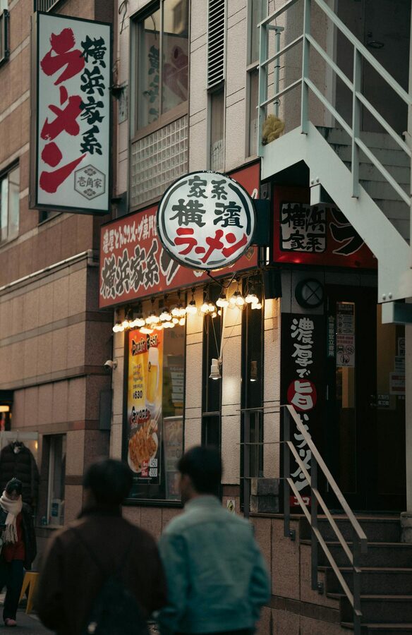 Two people walking past a traditional ramen shop in Japan at night