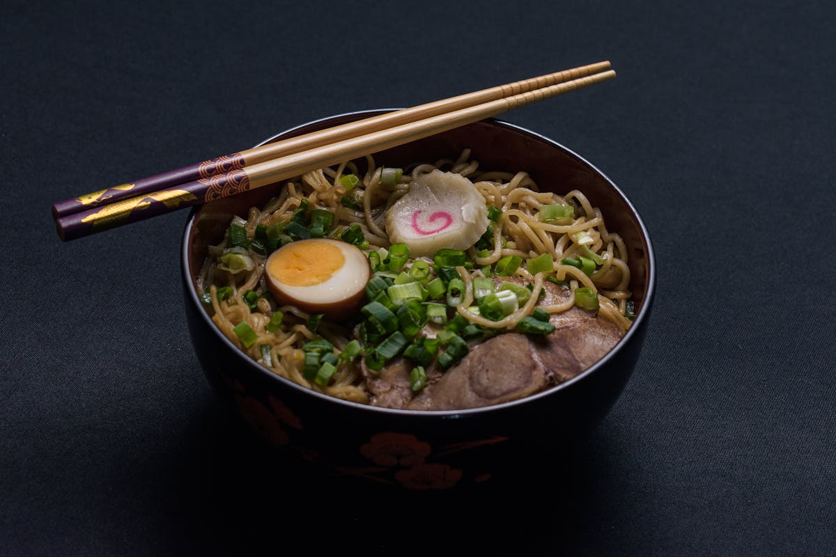 A steaming bowl of Japanese ramen topped with a halved soft-boiled egg, sliced scallions, and chopsticks resting on top