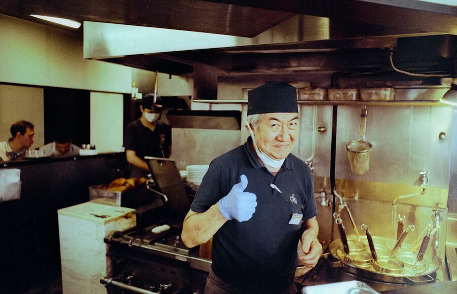 A cheerful chef giving a thumbs up gesture inside a traditional Japanese ramen restaurant kitchen