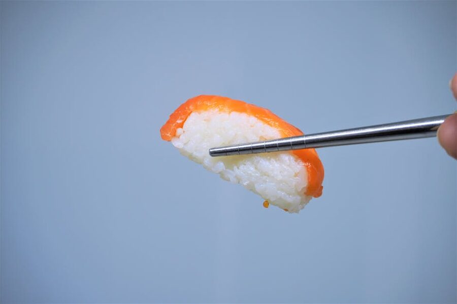 Piece of salmon sushi held with chopsticks against a plain backdrop