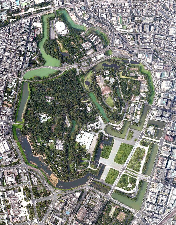 Aerial photograph of the Tokyo Imperial Palace grounds showing palace buildings surrounded by moats and green areas with downtown Tokyo beyond
